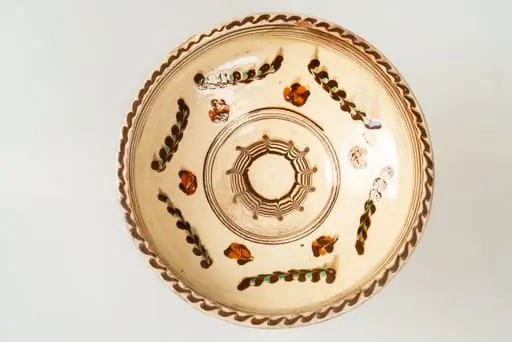 Farfurie adâncă, de tip castron, modelată la roata olarului, angobată pe fața interioară cu humă albă. Decorul este realizat prin pictare cu cornul și prin jirăvire: culorile sunt picurate cu cornul și trase (jirăvite) cu gaița. Motivele decorative sunt florale și vegetale stilizate și decor specific tehnicii prin jirăvire. Buza și bordura interioară a acesteia sunt marcate de linii concentrice sub formă de cerc. Muchia buzei este marcată de o linie continuă vălurită de culoare maro. Cromatica decorului: alb, verde, siena arsă, maro. Piesa este acoperită pe fața sa interioară de un strat de smalț transparent., by Unknown