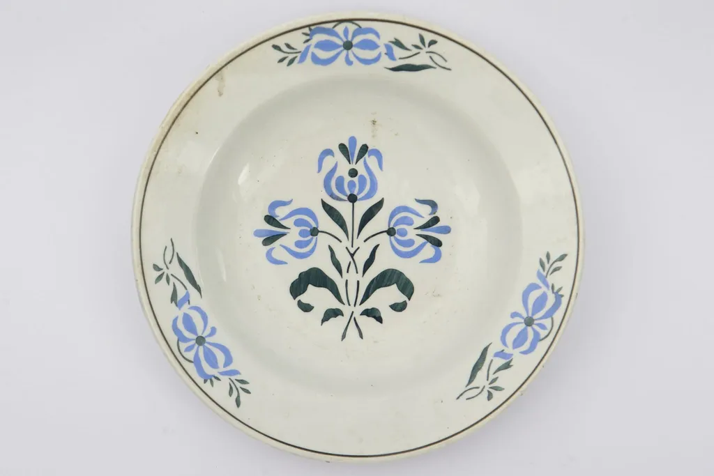 Farfurie cu fundul plat, puțin adâncit. Pe fundul vasului decor ilustrând un buchet de flori albastre cu tulpini și frunze verzi. Pe marginile vasului dispuse câte trei grupuri a două flori ca și cele de pe fund. La buză o linie neagră dispusă circular.. La posterior pe inelul de susținere practicate două perforații pentru atârnare. Cromatica: fond: alb; motive: albastru, verde, negru., by Unknown, 1850