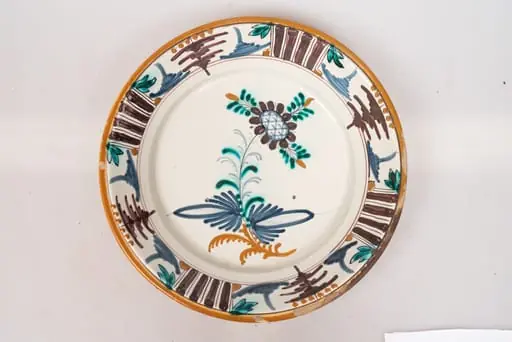 Farfurie de formă aplatizată, cu buza răsfrântă. Decorul, dispus pe un fond alb lăptos, prin pictare, este format dintr-un motiv floral stilizat, dispus central, pe fundul farfuriei, și motive geometrice (puncte galbene, grupuri de dungi paralele de culoare maro-brun, linii subțiri, paralele, linii sub formă de cerc, motivul bradului), dispuse într-un registru continuu pe buza farfuriei. Muchia buzei este marcată cu o dungă de culoare galbenă. Cromatica: alb, verde, brun, albastru, galben-ocru., by Unknown