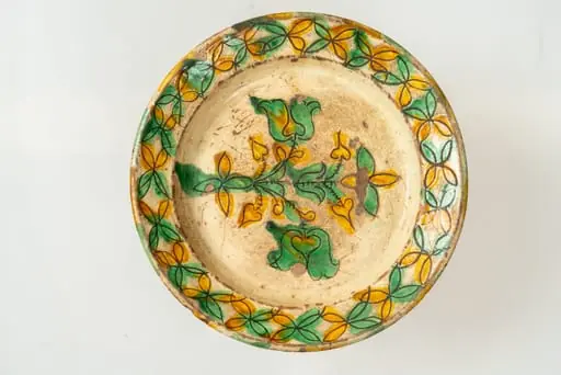 Farfurie de formă aplatizată, cu buză răsfrântă și muchie rotunjită. Piesa este modelată la roata olarului, angobată cu humă albă. Decorul este realizat prin sgrafitare în angoba crudă, peste care se aștern cu pensula culorile de smalț: verde și galben. Motivele decorative sunt florale și vegetale stilizate, dispuse pe fundul și pe buza farfuriei. Cromatica decorului: alb, galben, verde., by Unknown