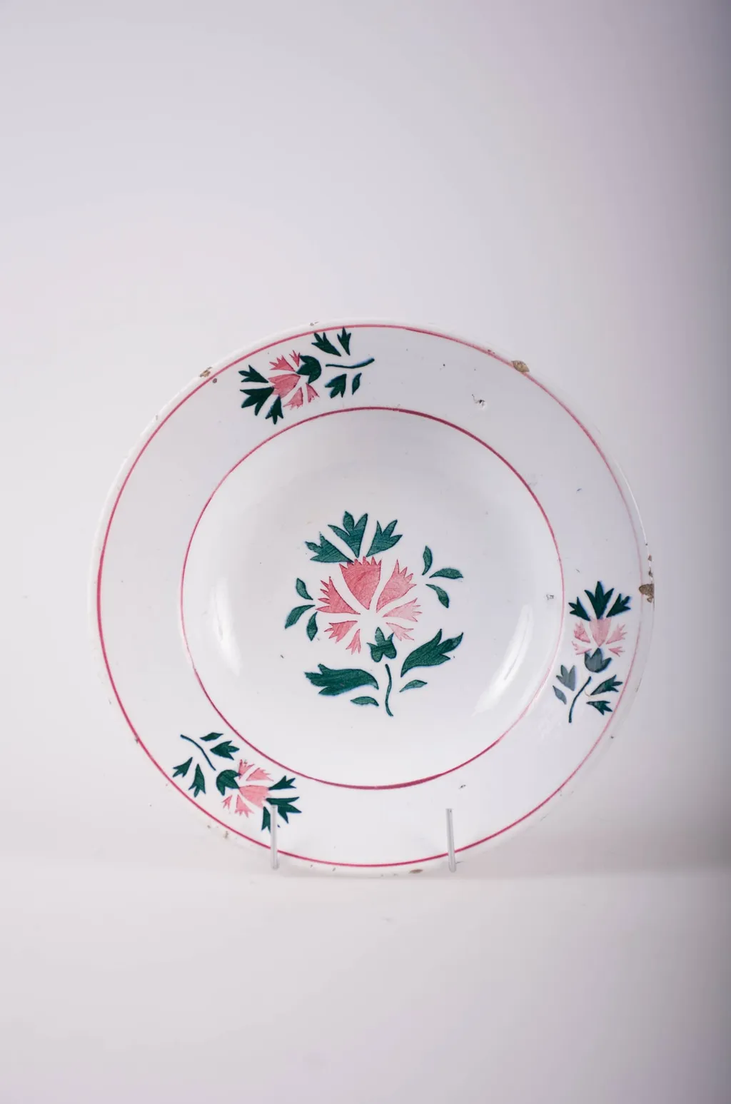 Farfurie de formă tronconică, adâncită; pe fund are găuri pentru agățare. În centru și pe margine este decorată cu motive fitomorfe și florale; pe margine sunt trei flori cu frunze încadrate de două cercuri concentrice; în interior este doar o floare cu frunze. Cromatică: fond: alb; motive: vișiniu; verde., by Unknown, 1850