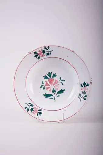 Farfurie de formă tronconică, adâncită; pe fund are găuri pentru agățare. În centru și pe margine este decorată cu motive fitomorfe și florale; pe margine sunt trei flori cu frunze încadrate de două cercuri concentrice; în interior este doar o floare cu frunze. Cromatică: fond: alb; motive: vișiniu; verde., by Unknown