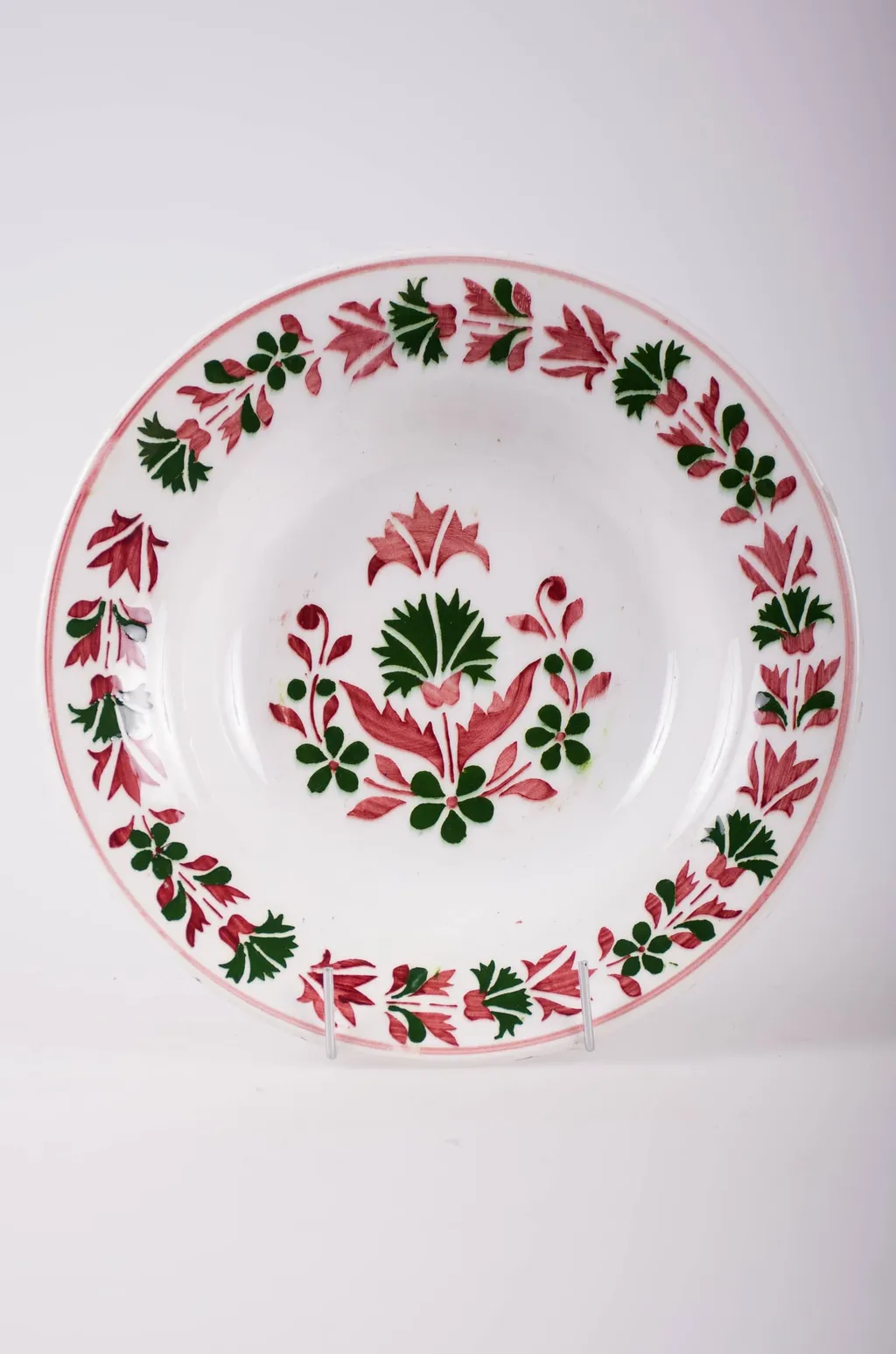 Farfurie de formă tronconică, adâncită; pe fund are găuri pentru agățare, Este decorată cu motive florale și geometrice; pe margine este decorat cu flori încadrate la exterior de o linie; în centru are mai multe flori. Pe fund are o marcă indescifrabilă. Cromatică: fond: alb; motive: vișiniu; verde., by Unknown, 1850