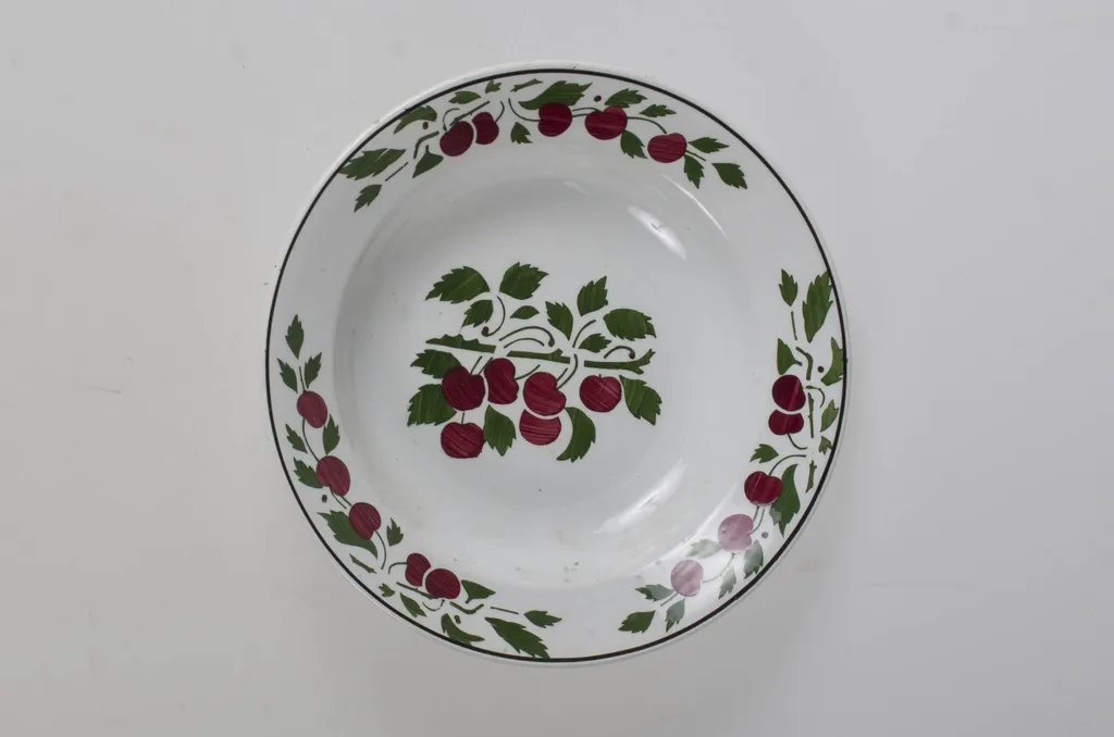 Farfurie de formă tronconică, adâncită; pe margine este decorat cu motive vegetale încadrate de o linie; în centru are o crenguță cu fructe (cireșe). Pe exterior la baza vasului prezintă trei cercuri concentrice și cifra ”26”. Cromatică: fond: alb; verde; roșu; negru. Pe fundul vasului sunt două incizii în care este introdus un șnur folosit la agățarea vasului pe perete., by Unknown, 1850