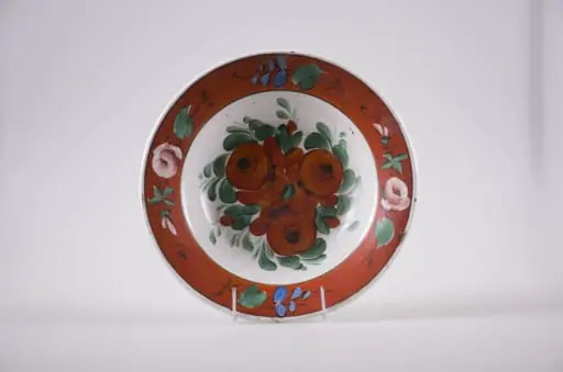Farfurie de formă tronconică din faianță, cu buza dreaptă, ornamentată prin pictare. Fundul este alb și are pictate trei flori roșii înconjurate de frunze mărunte, este delimitat de buza farfuriei cu o linie subțire, neagră. Buza vasului este roșie, decorată cu flori roz și frunzulițe discrete. În partea posterioară prezintă o gaură prin care se trece ața ce servește la agățarea farfuriei. Cromatică: fond: alb, roșu; motive: verde; roz; albastru; roșu; negru., by Unknown