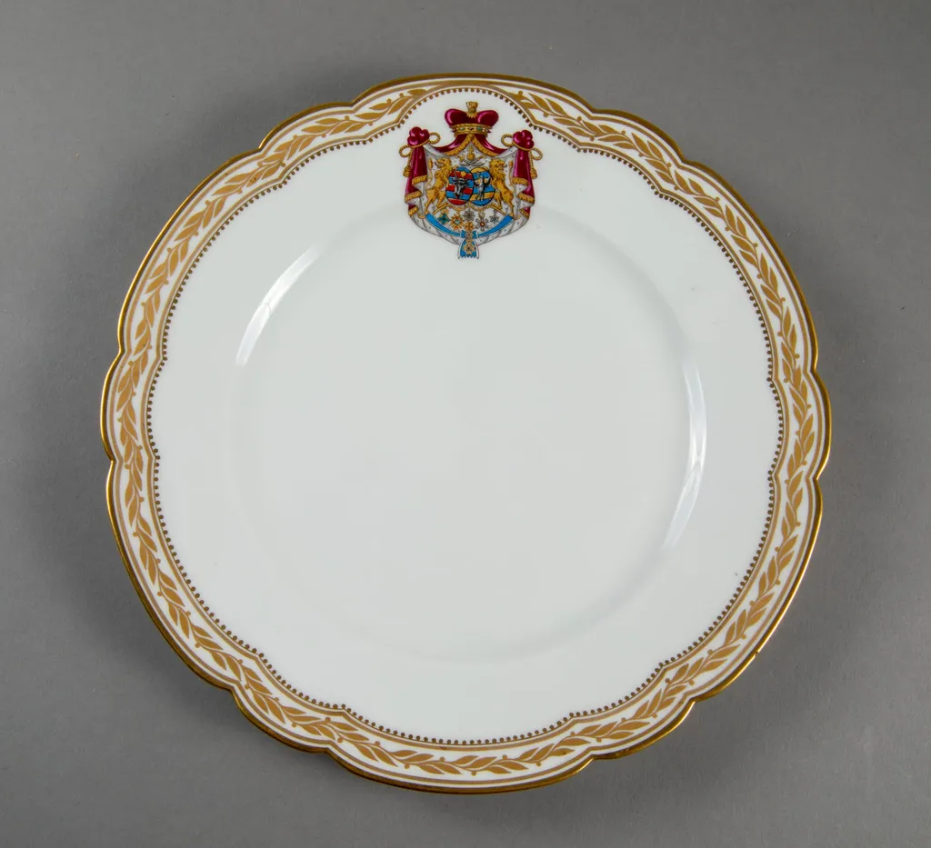 Farfurie de porțelan de Sevres, realizată din porțelan alb, cu margini treflate, decorate cu frunze de laur pictate, în centrul decorațiilor având blazonul familiei Suțu. Blazonul a fost adesea confundat cu cel al Principatelor Unite, fiind format din: stemele celor două țări românești: prima cea a Moldovei, cea de-a doua cea a Țării Românești, steme plasate pe două scuturi ovale, în fascie, acolate, puțin suprapuse, cea a Moldovei având capul de bour de argint cu stea în șase colțuri de aur, pe un scut cu segmente verticale egale roșii și albastre; cea a Țării Românești având acvila cruciată de argint, cu crucea de aur, scutul având segmente verticale galbene și albastre. Cele două scuturi au ca tenanți doi lei aurii, deasupra armele familiei Suțu – iataganul și sabia, ambele aurii, precum, pe un pavilion de purpură căptușit cu hermină, brodat cu ciucuri de aur și prins într-o coroană cu pietre scumpe. Pe scut, în partea inferioară se află și decorațiile familiei Suțu. Familia Suțu a fost o familie fanariotă grecească, venită în principate în cea de-a doua parte a secolului al XVIII-lea, având trei domni în ambele țări Românești (Alexandru Suțu, Mihai Suțu și Grigore Suțu). Blazonul familiei poate fi admirat inclusiv pe clădirea Palatului Suțu, astăzi Muzeul de Istorie a Municipiului București. Farfuria are marca atelierului Sevres și modelul din anul 1844, precum și seria., by Unknown, 1850