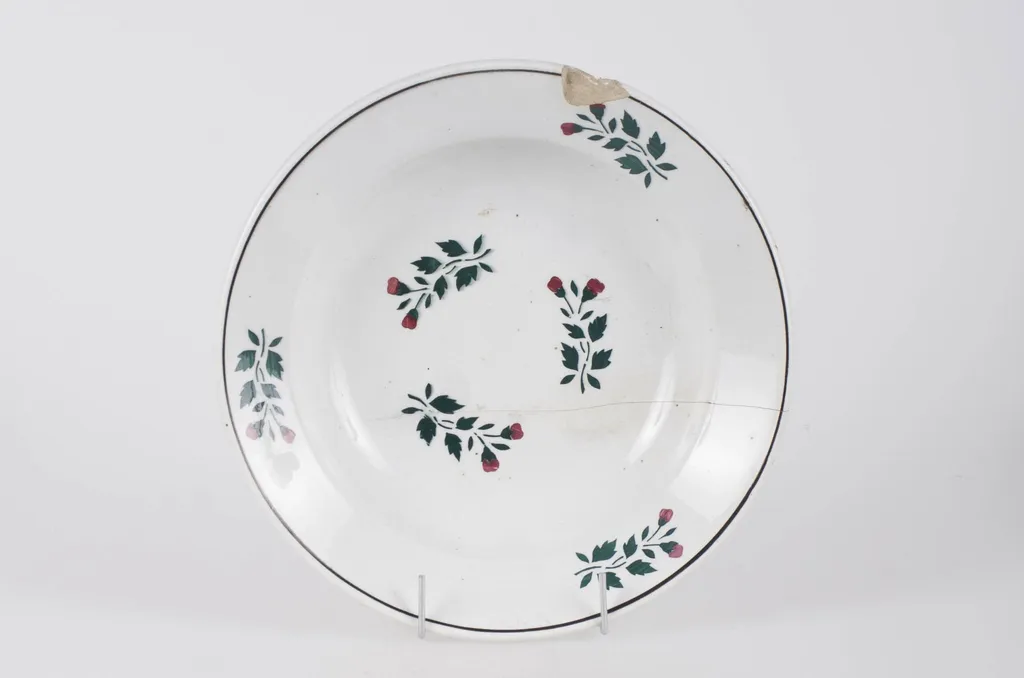 Farfurie din faianță smălțuită cu smalț alb. Vasul este de formă circulară, ușor adâncită. Pe fundul vasului decorul este floral-vegetal. Pe buza vasului se identifică trei flori, un decor geometric (cerc) ce încadrează decorul floral. Pe fundul vasului se identifică marca ”WILHELMSBURG”. Cromatică: fond alb; motive: verde; roșu., by Unknown, 1850