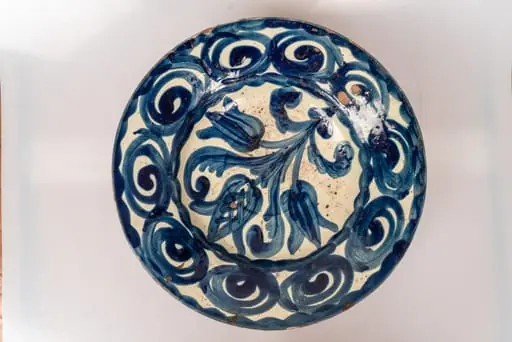 Farfurie mare, adâncă, cu buza răsfrântă; piesa este modelată la roata olarului și angobată pe fața interioară cu humă albă. Decorul monocromatic, este realizat prin pictare cu pensula, cu linii groase, și se compune din motive florale stilizate (trei flori de lalele cu frunze, dispuse central, pe fundul farfuriei) și motivul spiralei, ce acoperă întreaga suprafață a buzei, într-un șir continuu. Bordura interioară a farfuriei și muchia buzei sunt marcate de câte o linie vălurită. Pe fața ornamentată farfuria este acoperită cu un strat de smalț transparent. Cromatica: alb, albastru., by Unknown