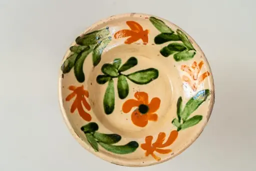 Farfurie mică, adâncă, cu buză răsfrântă. Piesa este modelată la roata olarului, angobată. Decorul este realizat prin pictare cu pensula și constă în motive decorative florale și vegetale (flori și frunze), dispuse central, pe fundul farfuriei și pe toată buza acesteia, sub formă de ghirlandă. Cromatica: alb, verde, siena naturală. Farfuria este smălțuită pe interior, cu smalț transparent., by Unknown