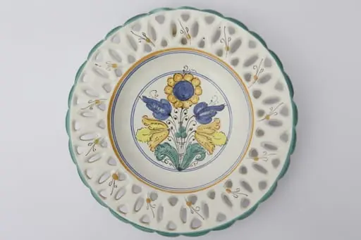 Farfurie ornamentală ce prezintă găurele între buză și partea adâncită obținute prin ștanțare. Buza este vălurită și prezintă un contur de culoare verde. Registrul central are motive florale și geometrice: cercuri galbene și albastre ce încadrează motivele florale. La posterior, baza inelară perforată pentru atârnare. Cromatică: fond: alb; motive: verde; galben; albastru; negru., by Unknown