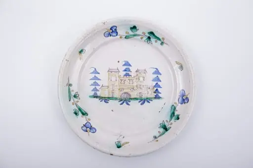 Farfurie plată,cu marginea lată și evazată spre interior. Marginea este decorată cu trei fire de floare de nu-mă-uita intercalate de un mic motiv fitomorf. Decorul central reprezintă un castel medieval poziționat într-un peisaj naturalist, înconjurat de trei brazi. Cromatică: alb, albastru, verde, maro., by Unknown