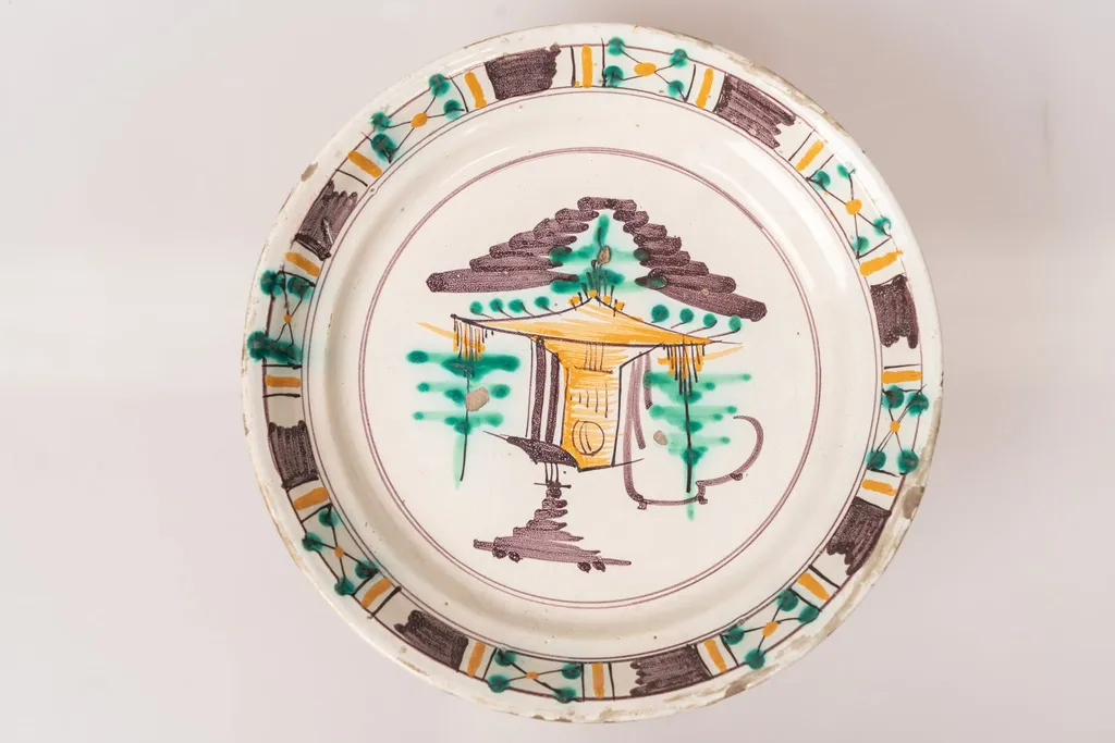 Farfurie semi-adâncă, cu buza răsfrântă. Decorul, dispus prin pictare pe un fond alb lăptos, este format dintr-un motiv arhitectural și motive vegetale stilizate, dispuse central, pe fundul farfuriei, și motive geometrice (linii galbene și dungi maro paralele, grupuri de linii intersectate sub formă de X, decorate cu puncte verzi și galbene), dispuse pe buza farfuriei, într-un registru continuu. Cele două registre ornamentale, cel central de pe fundul farfuriei și cel marginal, de pe buza acesteia, sunt încadrate în cercuri formate din linii fine de culoare maro-brun. Cromatica: alb, verde, maro-brun, galben ocru., by Unknown, 1850