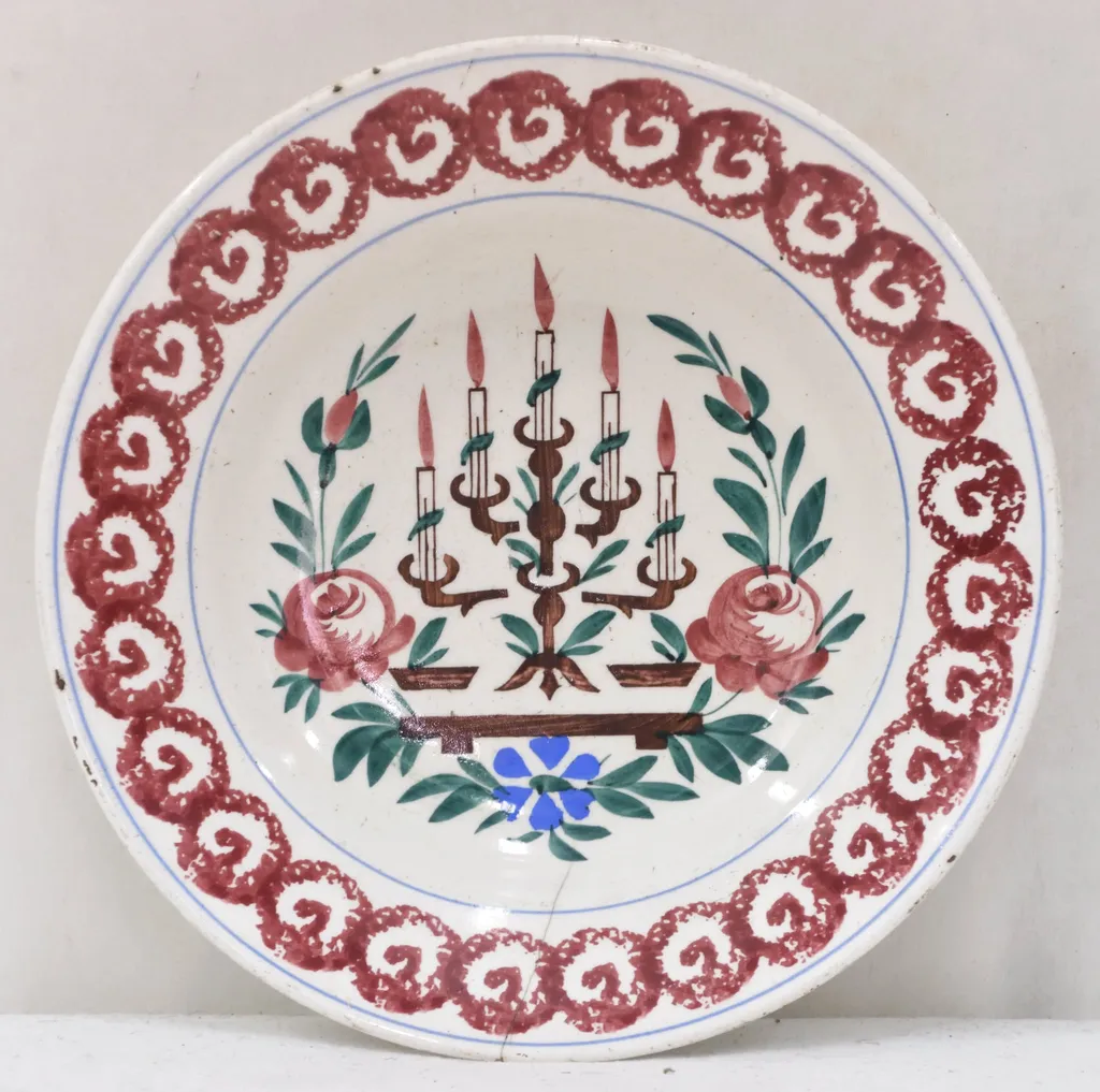 Obiectul are o formă tronconică cu baza mică în partea inferioară. Decorul se află numai la interiorul farfuriei fiind pictat cu motive geometrice, florale, vegetale și un simbol religios: pe fundul farfuriei a fost pictată o menoră cu cinci brațe (sfeșnic - obiect de cult evreiesc) sprijinită pe un suport; fiecare dintre brațele sfeșnicului susține câte o lumânare aprinsă; această reprezentare este încadrată de tulpini, frunze, flori și boboci (trei flori plus doi boboci); întreaga pictură de pe fundul farfuriei este încadrată de un cerc iar un altul este "trasat" de jur-împrejur în partea superioară, la buza piesei; între cele două cercuri se găsește un șirag format din cercuri (cu diametre egale) tangente la exterior, asemănător unui lanț cu zale rotunde; fiecare dintre aceste cercuri are la interior un semn, ce amintește de litera "V". Pe spatele farfuriei se află un mic orificiu rotund prin care se introduce firul textil pentru posibilitatea atârnării obiectului de perete. Culoarea piesei este albă iar pictura este realizată cu următoarele culori: maro închis, maro deschis, bleu și verde., by Unknown, 1850