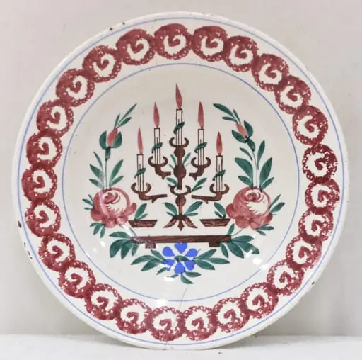Obiectul are o formă tronconică cu baza mică în partea inferioară. Decorul se află numai la interiorul farfuriei fiind pictat cu motive geometrice, florale, vegetale și un simbol religios: pe fundul farfuriei a fost pictată o menoră cu cinci brațe (sfeșnic - obiect de cult evreiesc) sprijinită pe un suport; fiecare dintre brațele sfeșnicului susține câte o lumânare aprinsă; această reprezentare este încadrată de tulpini, frunze, flori și boboci (trei flori plus doi boboci); întreaga pictură de pe fundul farfuriei este încadrată de un cerc iar un altul este "trasat" de jur-împrejur în partea superioară, la buza piesei; între cele două cercuri se găsește un șirag format din cercuri (cu diametre egale) tangente la exterior, asemănător unui lanț cu zale rotunde; fiecare dintre aceste cercuri are la interior un semn, ce amintește de litera "V". Pe spatele farfuriei se află un mic orificiu rotund prin care se introduce firul textil pentru posibilitatea atârnării obiectului de perete. Culoarea piesei este albă iar pictura este realizată cu următoarele culori: maro închis, maro deschis, bleu și verde., by Unknown