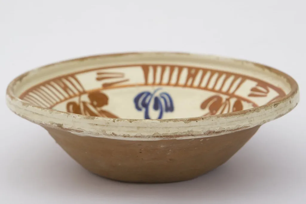 Piesă ceramică de dimensiuni medii, smălțuit la interior, având formă tronconică, cu baza plată, pereții ridicați și buza evazată. Întreaga suprafață interioră a piesei a fost decorată cu motive geometrizante și fitomorfe stilizate. O floare, cu petale verzi, roșii și albastre și cu un punct roșu în mijloc, este mărginită de un brâu circular, compus din opt reprezentări florale, cu aceiași cromatică. Marginal, pe buza piesei a fost realizat cel de-al doilea registru: două linii circulare, care încadrează patru registre, compuse din linii verticale, despărțite de alte patru registre din linii orizontale (2-3 linii). Farfuria era folosită în gospodărie pentru decorarea interioarelor. Prezintă uzură funcțională. Cromatică: fond: cărămiziu (exterior); crem (interior); motive: roșu; albastru; verde., by Unknown, unspecified, 1850