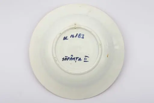 Vas ceramic de dimensiuni reduse, având formă tronconică, cu baza plată, pereții reficați și marginile evazate. Peste fondul alb au fost realizate decorațiile geometrizante și fitomorfe stilizate, dispuse în două registre. Primul registru este format dintr-un trandafir portocaliu cu frunze verzi, având în laterale câte o floare cu mijlocul albastru și petalele roșii, și câte un boboc roșu. Al doilea registru este format dintr-o tulpină circulară verde, pe care se dezvoltă trei grupuri de flori mici albastre. Alte trei grupuri, a câte trei linii portocalii oblice, însoțesc decorul fitomorf. Acest ultim registru este încadra dinspre interior de bandă groasă portocalie. Buza farfuriei a fost la rândul ei conturată cu portocaliu. Prezintă uzură funcțională. Cromatică: fond: alb; motive; portocaliu; verde; albastru., by Unknown