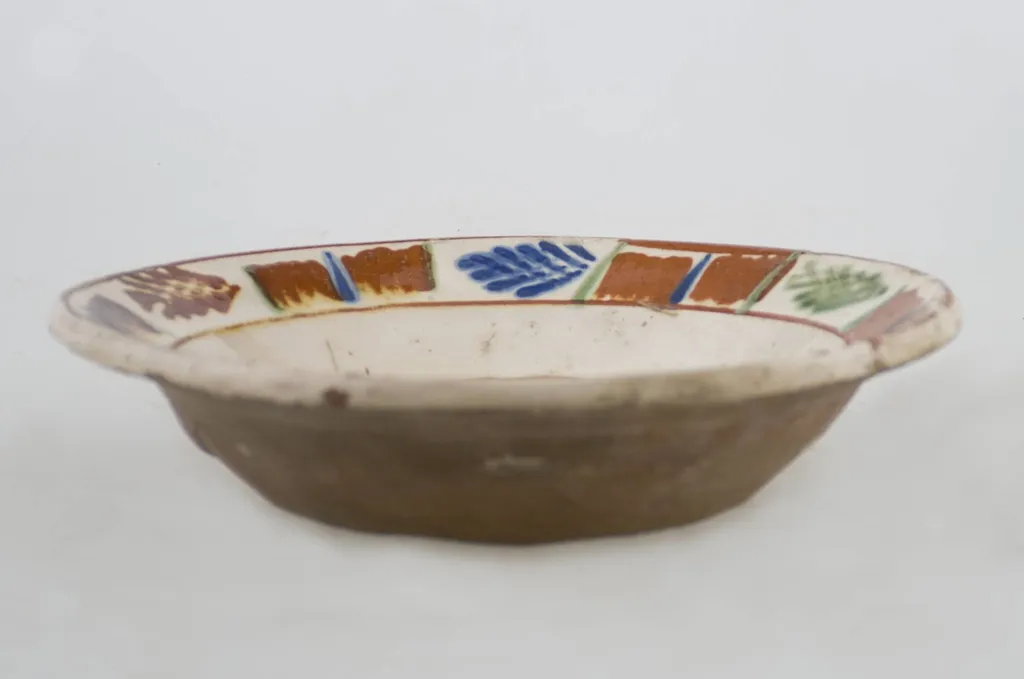 Vas ceramic de formă tronconică baza plată, pereții larg evazați și buza profilată. După modelarea la roată, uscarea și arderea recipientului, a fost aplicat un strat de angobă în interiorul vasului, peste care a fost pictat decorul floral și vegetal stilizat, format din două registre. În primul registru, situat pe fundul vasului și încadrat de o linie circulară, au fost realizate trei flori, cu petale albastre și mijlocul maro-roșcat, în jurul cărora se regăsesc petale verzi. Cel de-al doilea registru, cel care decorează buza vasului și este încadrat la rândul lui între două linii circulare, a fost decorat cu frunze și pete, despărțite de linii verticale. Cromatică: fond: alb; motive: maro-roșcat; albastru; verde., by Unknown, unspecified, 1850