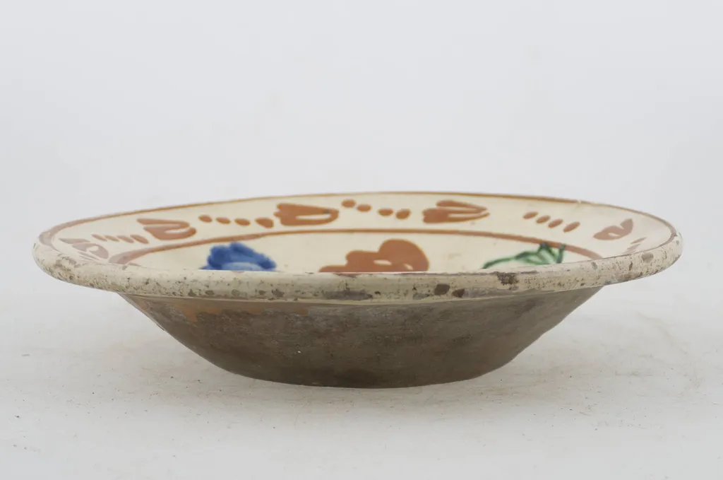 Vas ceramic de formă tronconică cu baza plată, pereții larg evazați și buza profilată. După modelarea la roată, uscarea și arderea recipientului, a fost aplicat un strat de angobă în interiorul vasului, peste care a fost pictat decorul floral și vegetal stilizat, format din două registre. Primul registru, situat pe fundul și pereții vasului, și încadrat de o linie circulară, a fost decorat cu motive florale stilizate: pe fundul vasului se află o floare, cu petalele de diferite culori. Pereții recipientului ceramic au fost decorați la rândul lor cu opt flori, dispuse concentric. Al doilea registru, cel care decorează buza recipientului ceramic, și este mărginit la rândul lui de o linie circulară, prezintă un brâu compus din nouă flori cu tulpinile formate din patru puncte. Cromatică: alb la interior; cărămiziu la exterior; motive: albastru; verde; maro-roșcat., by Unknown, unspecified, 1850