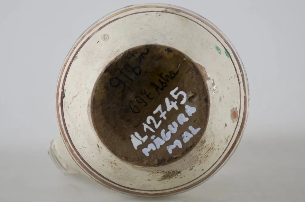 Vas ceramic smălțuit de mici dimensiuni, piriform, cu baza plată și bombat în zona „pântecelui”. Prezintă o toartă ce pornește din zona gâtului redus ca dimensiuni și se termină în zona diametrul maxim al vasului. Decorul vasului este compus din trei registre picturale. În primul registru au fost pictate două flori albe pe fundal verde și o a treia pe fundal maro, fiind despărțite prin linii de culoare galbenă. Cel de-al doilea registru a fost decorat cu motive geometrice stilizate, linii și triunghiuri afrontate de culoare galbenă, pe fundal de culoare verde. Ultimul registru a fost decorat cu linii de culoare maro și galben, asemenea cu trei brazi și înconjurate de câte două frunze de culoare verde. Toate cele trei registre picturale sunt încadrate de câte trei linii concentrice (două subțiri și una lată) de culoare maro-închis și maro-roșcat. Pe toartă este decorat cu linii paralele orizontale. Cromatică: fond alb; motive: galben; verde; maro., by Unknown, unspecified, 1850
