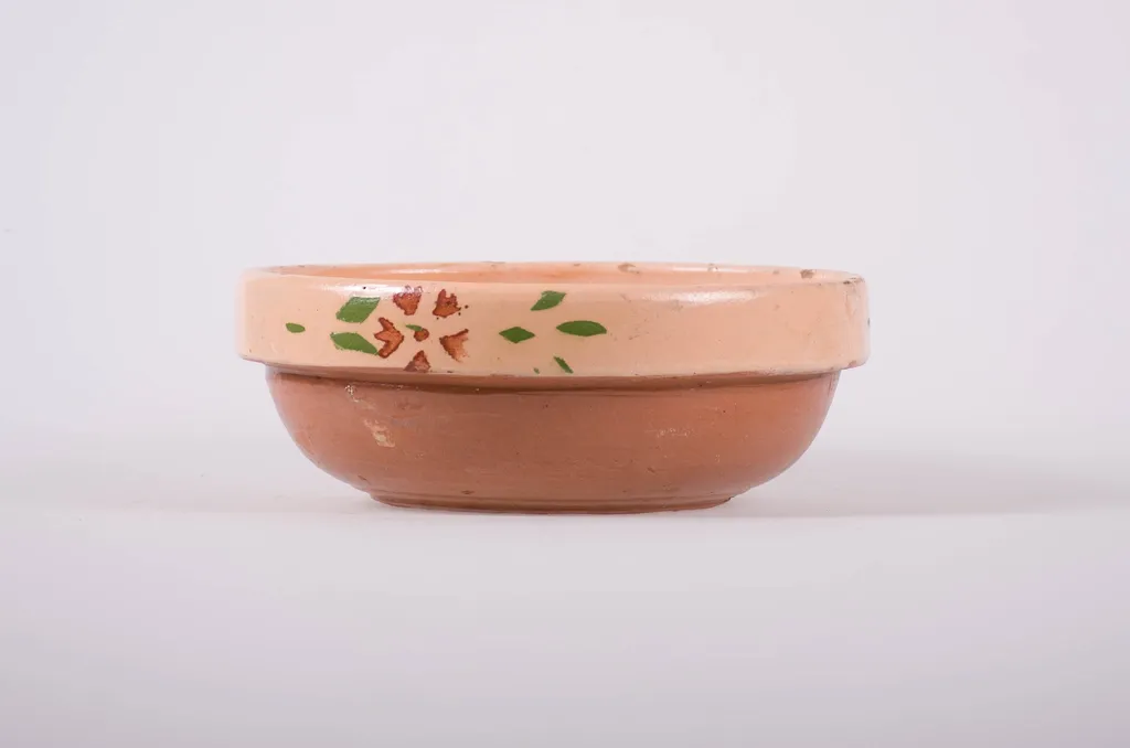Vas de formă tronconică, adâncit, cu buza foarte înaltă; pe fund are găuri pentru agățare. Este smălțuit interior și exterior. Decorat cu motive geometrice: linie; motive florale. Cromatică: fond; crem; motive: maron; verde., by Unknown, 1850