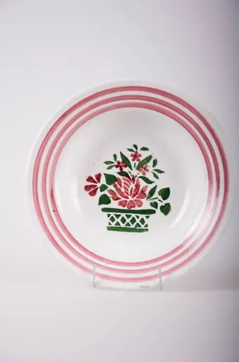 Vas de formă tronconică, adâncit; pe fund are găuri pentru agățare. În centru este decorată cu motive florale: vază cu flori; pe margine sunt trei cercuri concentrice; motive geometrice: linie, romb, triunghi. Pe fund are o marcă indescifrabilă. Cromatică: fond: alb; motive: vișiniu; verde., by Unknown