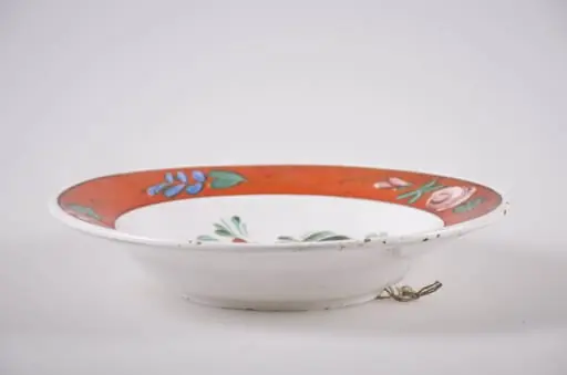 Vas de formă tronconică din faianță, cu buza dreaptă, ornamentată prin pictare. Fundul alb, pe care sunt pictate trei flori roșii (bujor) înconjurate de frunze mărunte, este delimitat de buza farfuriei cu o linie subțire, neagră. Buza vasului este roșie, decorată cu flori roz și frunzulițe discrete verzi și albastre. În partea posterioară prezintă o gaură prin care se trece ața ce servește la agățarea farfuriei. Cromatică: fond: alb; motive: roșu; verde; roz; albastru; negru., by Unknown