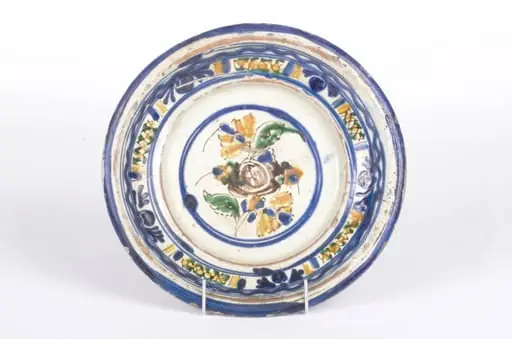 Vasul ceramic este de formă circulară, ușor adâncit, cu pereți evazați și ondulați. Decorul este amplasat pe două registre: primul registru se găsește la partea inferioară a buzei și se înscrie perimetral pe aceasta cu motive geometrice și florale (cercuri), al doilea registru este plasat central pe fundul vasului și prezintă același tip de motive. Cromatică: fond: alb-crem; motive: albastru; galben; verde; maro., by Unknown