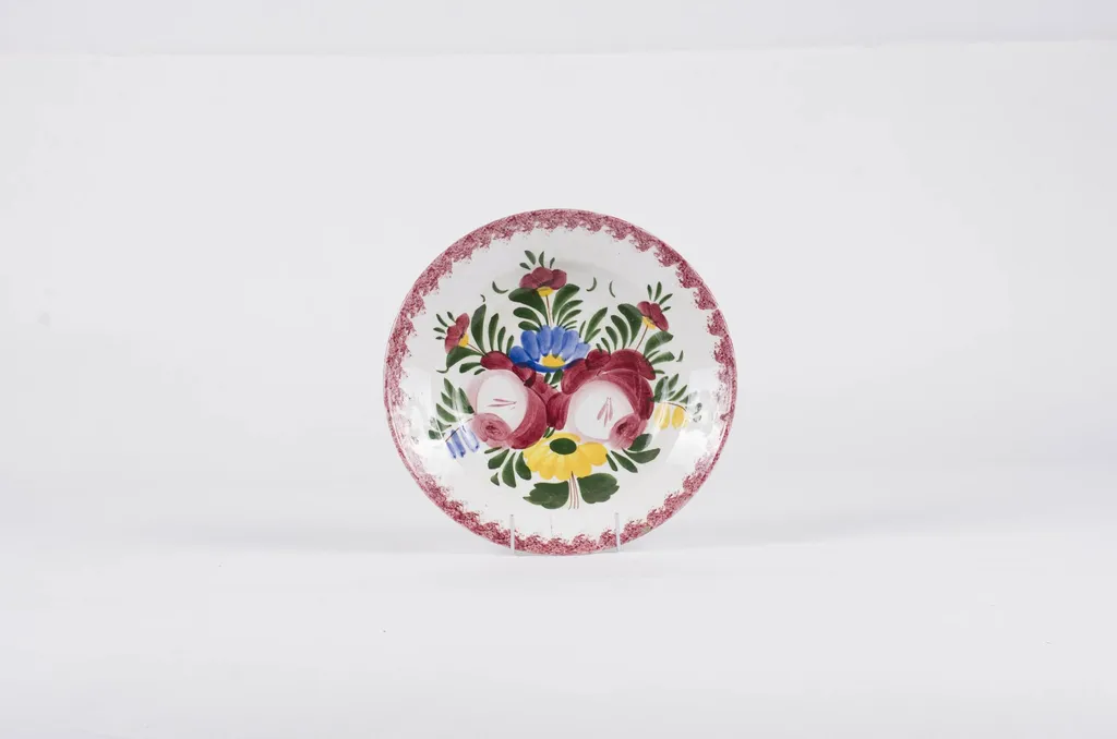Vasul este de formă tronconică fiind smălțuit interior-exterior. Este confecționat din faianță Decorul este format din motive florale pictate pe fond alb; gura este decorată în stropi de culoare vișinie iar baza farfuriei este decorată cu motive florale. Se identifică șapte flori alcătuind motivul decorativ pe baza farfuriei. Pe spate inscripția este indescifrabilă. Cromatică: galben; albastru; verde; vișiniu; alb;, by Unknown, 1850