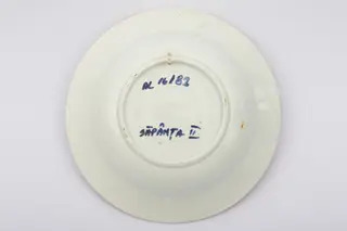 Vas ceramic de dimensiuni reduse, având formă tronconică, cu baza plată, pereții reficați și marginile evazate. Peste fondul alb au fost realizate decorațiile geometrizante și fitomorfe stilizate, dispuse în două registre. Primul registru este format dintr-un trandafir portocaliu cu frunze verzi, având în laterale câte o floare cu mijlocul albastru și petalele roșii, și câte un boboc roșu. Al doilea registru este format dintr-o tulpină circulară verde, pe care se dezvoltă trei grupuri de flori mici albastre. Alte trei grupuri, a câte trei linii portocalii oblice, însoțesc decorul fitomorf. Acest ultim registru este încadra dinspre interior de bandă groasă portocalie. Buza farfuriei a fost la rândul ei conturată cu portocaliu. Prezintă uzură funcțională. Cromatică: fond: alb; motive; portocaliu; verde; albastru.