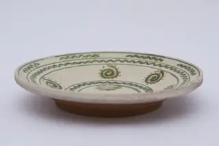 Vas ceramic de formă discoidală pe bază puțin înălțată. Fața farfuriei a fost introdusă în angobă albă, decorația a fost făcută cu pensula. Pe fundul vasului sunt două cercuri concentrice: în primul sunt doi brăduți, iar în al doilea valul punctat. Pe pereți sunt șase spirale punctate. Marginea vasului a fost împărțită în două registre, primul cu val punctat, în al doilea sunt șapte valuri întrerupte. La posterior, pe fund, semătura ”D Șchiopu ''. Pentru atârnare baza este perforată. Cromatică: fond: alb; motive; verde; galben.