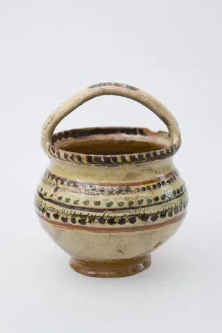 Vas ceramic de mici dimensiuni, pe bază inelară, pântecos, cu fundul și gura late. Deasupra gurii vasului a fost pusă o toartă de prindere. Buza vasului a fost pictată printr-un brâu de crenguțe. Pe pântecul vasului brâuri de puncte și două benzi subțiri jirăvite. Smălțuit în interior și exterior. Cromatică: fond: crem; motive: verde; maro; bej., 1850
