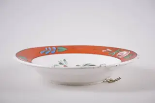 Vas de formă tronconică din faianță, cu buza dreaptă, ornamentată prin pictare. Fundul alb, pe care sunt pictate trei flori roșii (bujor) înconjurate de frunze mărunte, este delimitat de buza farfuriei cu o linie subțire, neagră. Buza vasului este roșie, decorată cu flori roz și frunzulițe discrete verzi și albastre. În partea posterioară prezintă o gaură prin care se trece ața ce servește la agățarea farfuriei. Cromatică: fond: alb; motive: roșu; verde; roz; albastru; negru., 1850