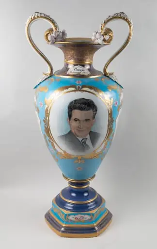 Vază din porțelan, fond albastru, tip amforă, cu anse, decorată cu două medalioane, unul reprezentând portret Nicolae Ceaușescu, cel de-al doilea un buchet cu flori. Pe gâtul vazei o dedicație ”Omagiu, Ianuarie 1989”. Vază oferită lui Nicolae Ceaușesacu de Consiliul Național al Femeilor cu ocazia aniversării zilei de naștere, ianuarie 1989.